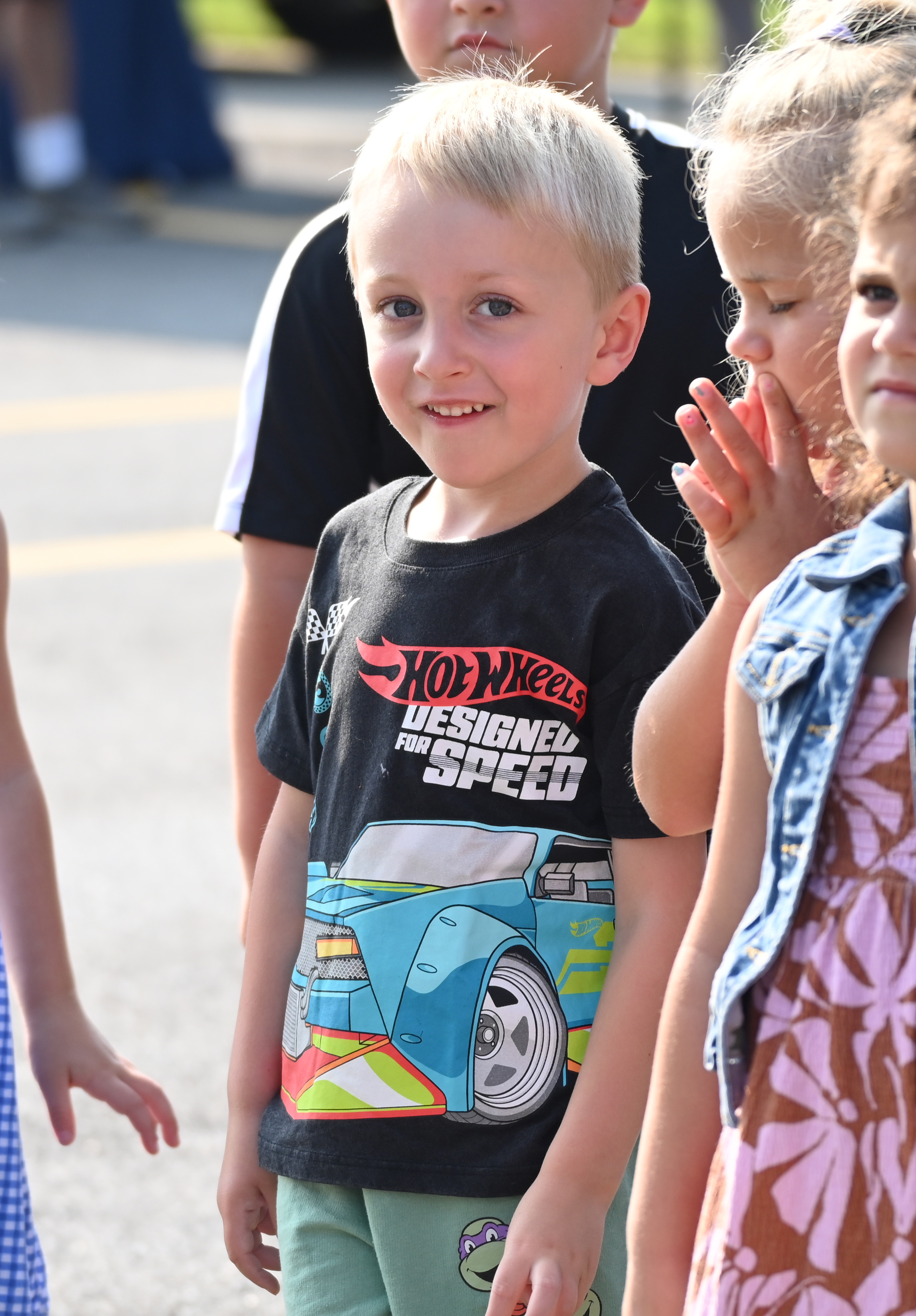 A young kindergarten boy in a black Hot Wheels t-shirt smiles while standing with other children.